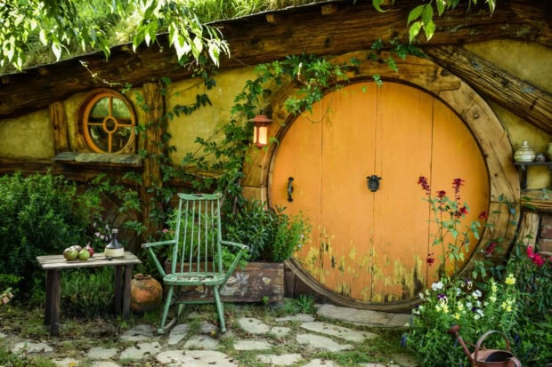 A charming hobbit house with a round wooden door and a green chair outside surrounded by plants and flowers