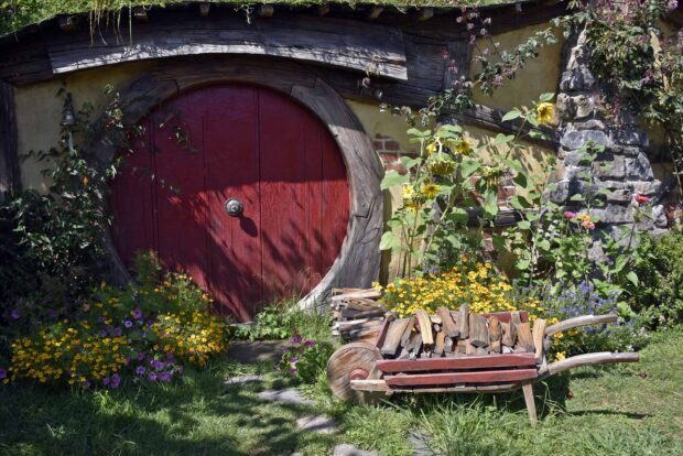 A charming hobbiton door surrounded by colorful flowers and a wooden wheelbarrow filled with firewood