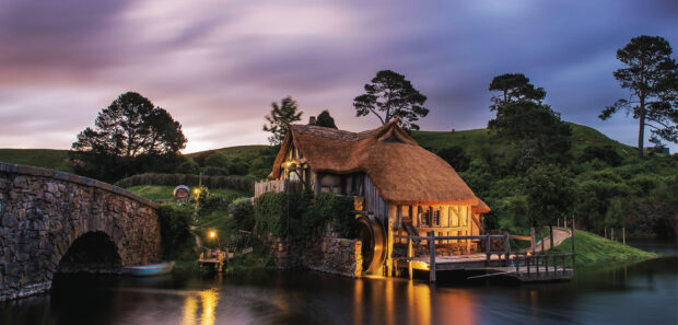 A peaceful watermill in Hobbiton surrounded by green hills and trees at twilight