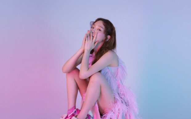 A K Pop singer wearing a pink feather outfit sitting with her hands near her face
