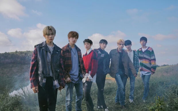 A K Pop group posing outdoors on a grassy hill under a blue sky