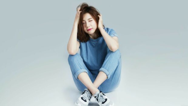 Young K Pop star sitting cross legged in denim outfit and sneakers winking at camera