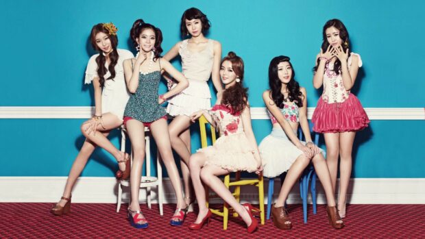 A K Pop girl group posing stylishly against a blue wall with vibrant outfits and high heels