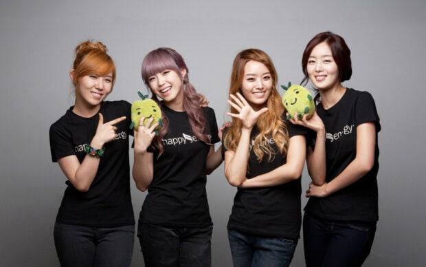 Four K pop members smiling and posing with green plush toys in a studio setting