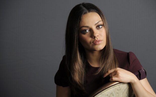 Mila Kunis wearing a burgundy dress sitting on a chair with a thoughtful expression