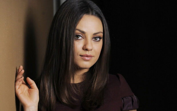 Mila Kunis with long dark hair posing against a wall in a dark outfit