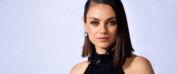 Mila Kunis with short hair wearing a black outfit and earrings on a light background