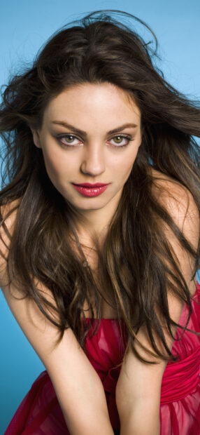 Mila Kunis wearing a red dress with flowing hair against a blue background