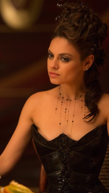 Mila Kunis wearing an elegant black dress with intricate necklace in a dimly lit setting