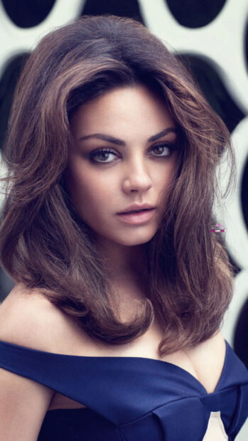 Mila Kunis with long brown hair wearing a blue off shoulder dress looking at the camera