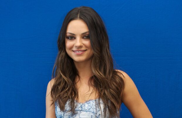 Mila Kunis with long wavy hair smiling in front of a blue background