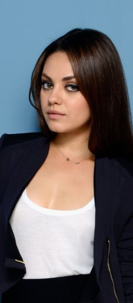 Mila Kunis wearing a white top and dark jacket posing against a blue background