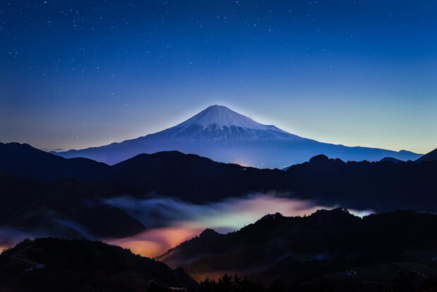 Mount Fuji Japan seen under a starry night with colorful fog over dark hills