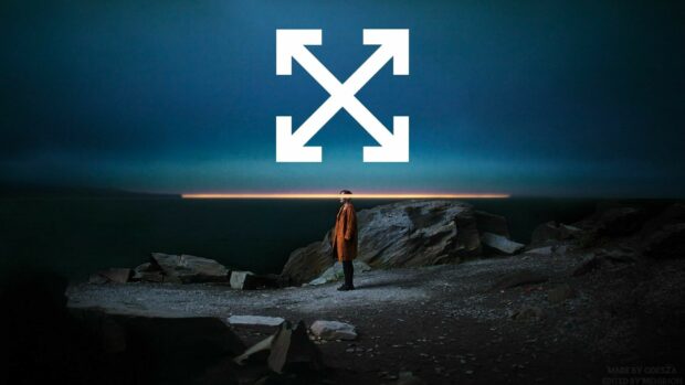 A person in an off white coat stands on rocky terrain near the ocean at dusk