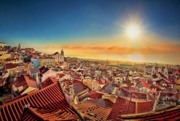 A panoramic view of Portugal cityscape with red rooftops and a bright sun over the ocean at sunset