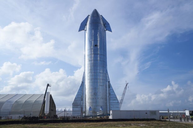 A shiny starship standing upright on a launch site under a partly cloudy sky