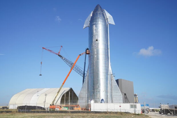 Large metal starship under construction at launch site with cranes and blue sky background