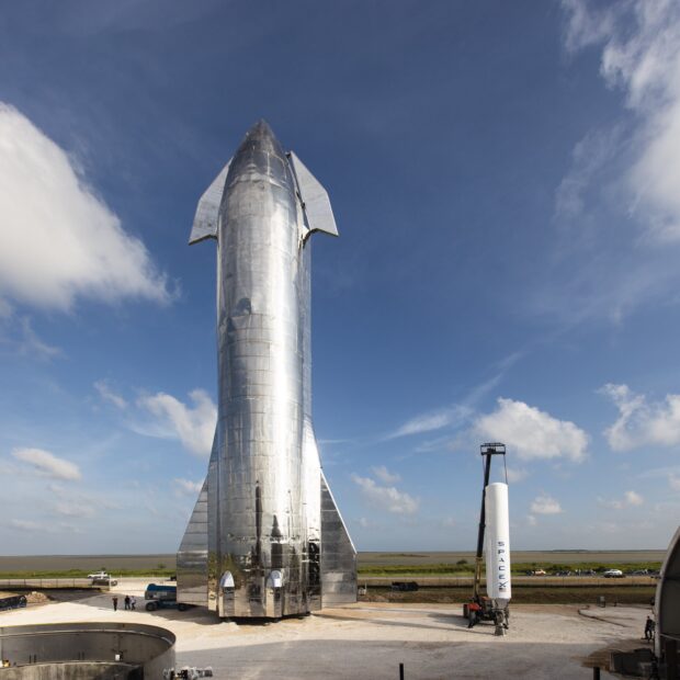 A shiny starship standing vertically on a launch pad under a clear blue sky