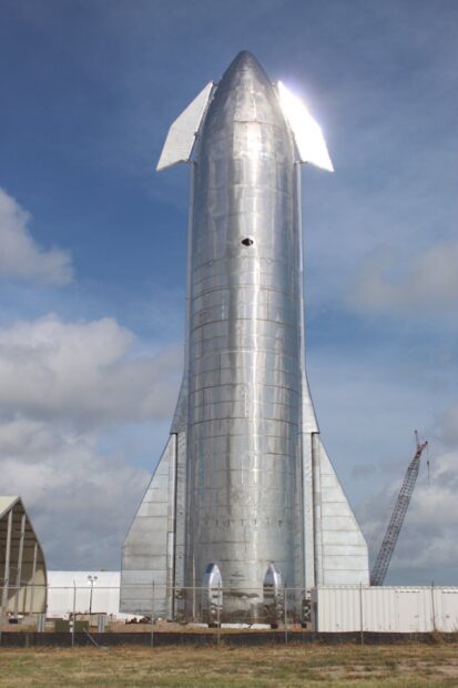 A shiny metal starship standing vertically on a launch site under a partly cloudy sky