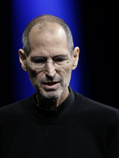 Steve Jobs with glasses and black turtleneck speaking on stage with blue background
