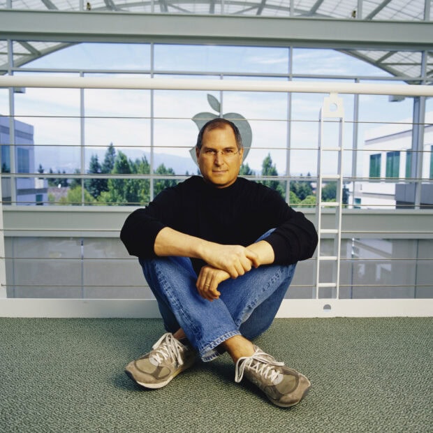Steve Jobs sitting cross legged wearing a black turtleneck and jeans in front of an Apple logo