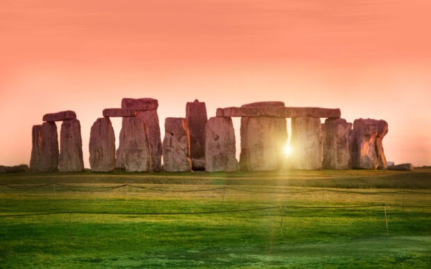 Ancient Stonehenge monument with sun shining through stones at sunset on green field