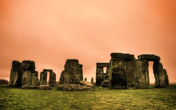 Ancient Stonehenge monument under an orange sky with green grass in the foreground