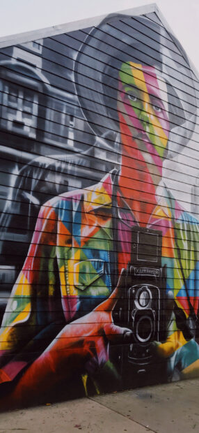 A colorful street art painting of a person holding a vintage camera on a wooden wall