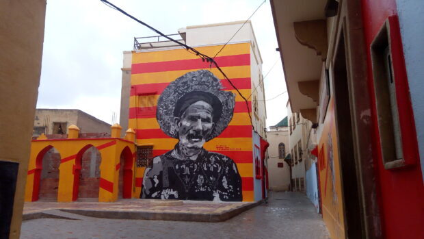 A black and white street art mural of a man wearing a hat on a colorful building wall