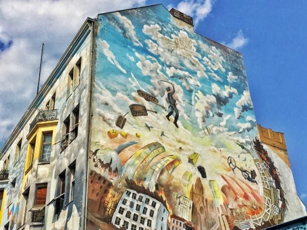 A detailed street art mural depicting a surreal scene with tanks flying in the sky and burning buildings below
