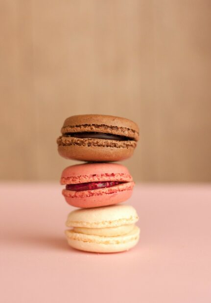 A stack of colorful macarons with chocolate raspberry and vanilla flavors on a soft pink surface