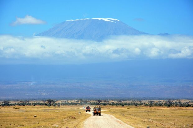 Vehicles traveling on a dirt road with Kilimanjaro mountain in Tanzania in the background surrounded by clouds