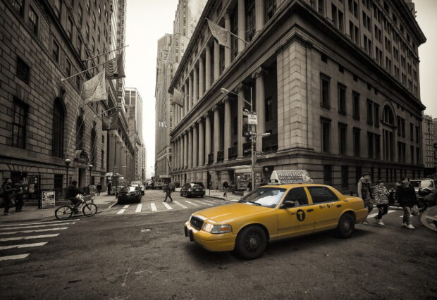 A yellow taxi driving through a busy city street with tall buildings and pedestrians crossing the road