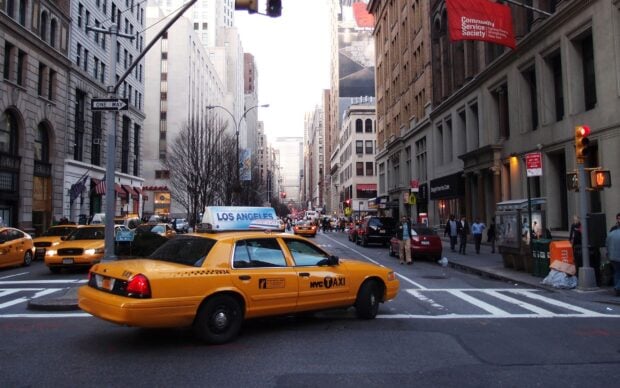 Yellow NYC taxi driving through busy city street with tall buildings and pedestrians