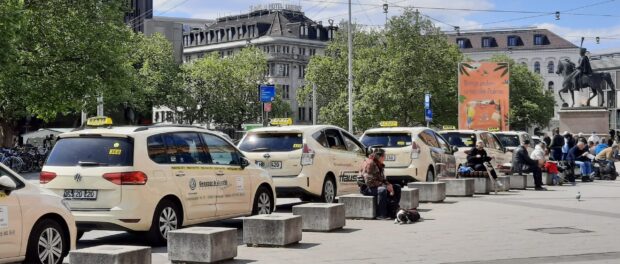 A row of taxi vehicles parked along a city street with people sitting on nearby benches and trees in the background