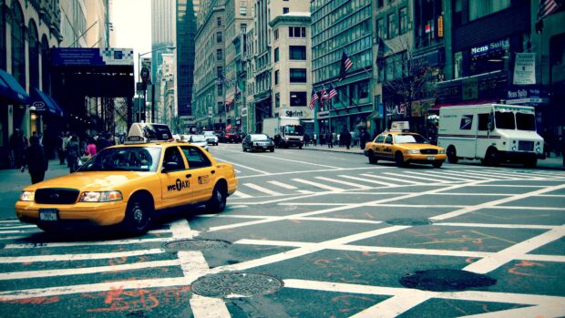 Yellow taxi driving on a busy city street with tall buildings in the background