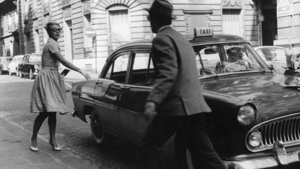 A vintage taxi with a driver and a woman in a striped dress hailing the taxi on the street