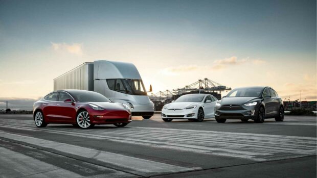 A lineup of Tesla Model Y vehicles and a semi truck parked on an industrial pier at sunset