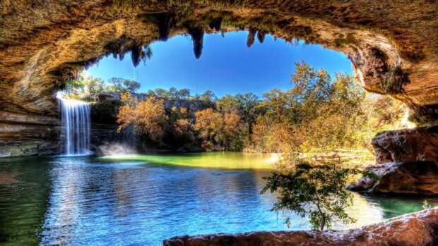 A serene Texas Hill Country scene featuring a waterfall and lush trees viewed from a cave opening