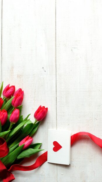A bouquet of red tulips with a red ribbon and a love card on a white wooden table