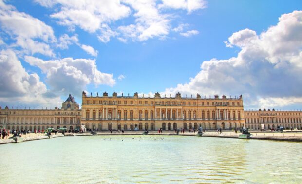 The grand Versailles palace with a large reflecting pool under a bright blue sky