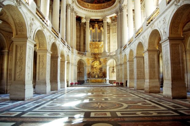 The grand hall of Versailles palace features intricate marble floors and detailed stone columns with golden decorations