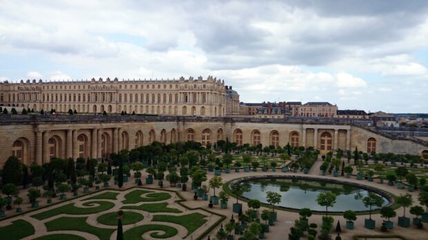The Versailles palace with formal gardens and a large reflective pond under a cloudy sky