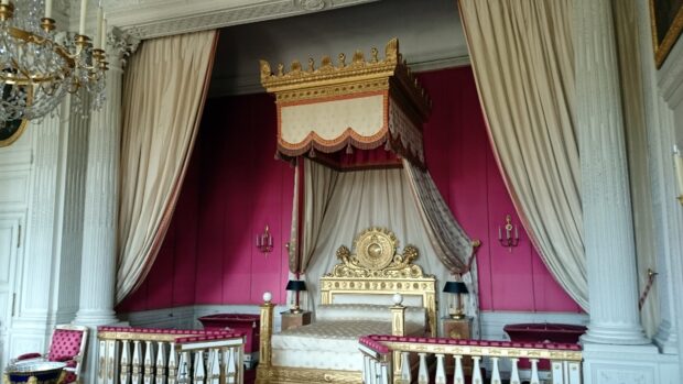 A luxurious Versailles room featuring a gold ornate bed with white and pink drapery and decorated walls