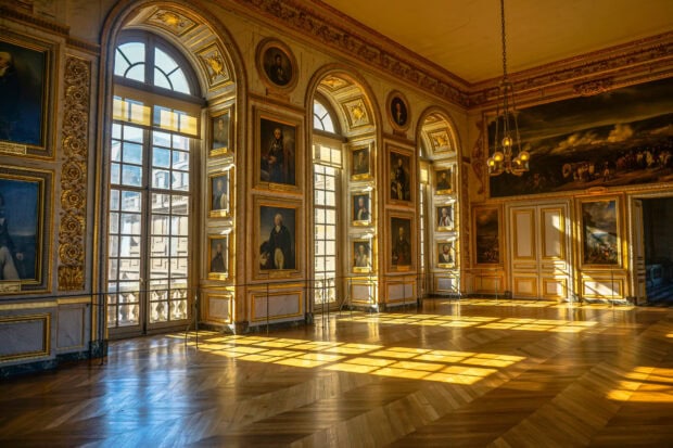 Interior view of a Versailles hall with historical portraits and sunlight streaming through large windows