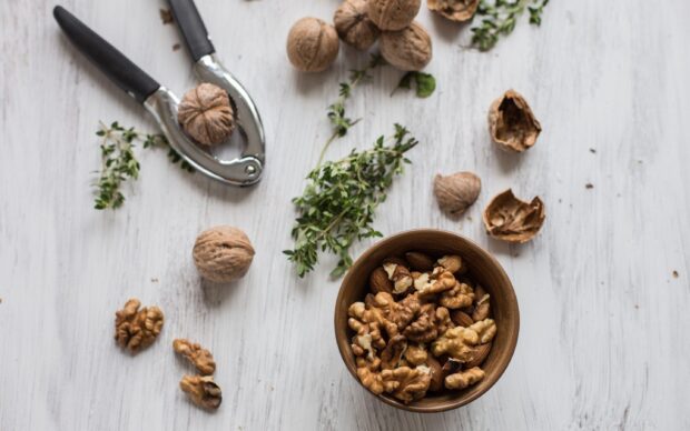A wooden bowl filled with walnuts and almonds surrounded by whole walnuts and nutcracker on a white wooden surface