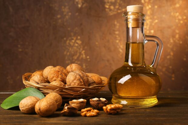 A basket of walnuts with a glass bottle of walnut oil on a wooden surface and a green leaf nearby