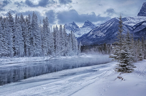 Snowy forest and frozen river in winter with mountain peaks in the background