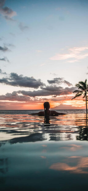 Woman enjoying summer sunset in pool with ocean view and palm tree