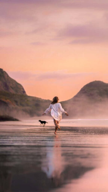 Woman enjoying a summer evening run on the beach with a dog and mountains in the background